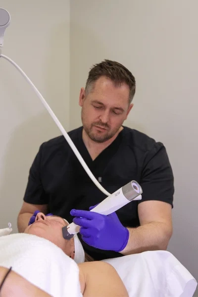 A close-up of a technician's hands with blue gloves using an aesthetic device with a metal tip on a client's cheek.
